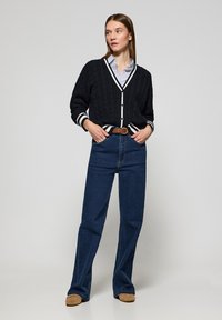 Marinblå kabelstickad cardigan med vit kant, svarta knappar, lagd över en ljusblå randig skjorta; kombinerad med högmidjade mörka denimjeans.