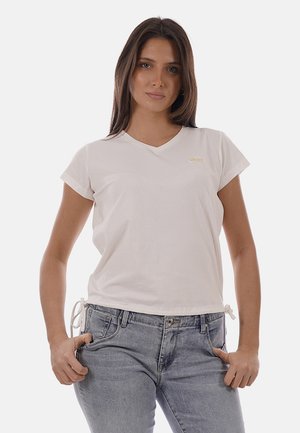 Donna con capelli lunghi e scuri che indossa una maglietta bianca a maniche corte con scollo a V e jeans azzurri, in piedi con le mani nelle tasche.