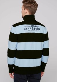 Maglione a righe neri e azzurri chiari con colletto alto. La parte posteriore presenta un logo con la scritta: "Coastal Waters Camp David" in lettere scure.