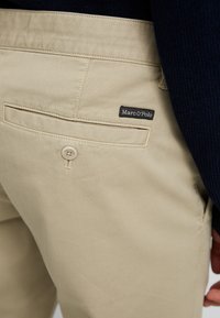 Beige cotton trousers featuring a back pocket with a button closure and a black fabric label reading "Marc O'Polo." Smooth texture.