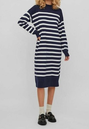 Frau trägt ein langärmeliges Midikleid mit horizontalen Streifen in Marineblau und Weiß, cremefarbene Socken und schwarze Loafers, stehend vor einem einfarbigen Hintergrund.