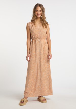 Frau lächelnd, trägt ein ärmelloses, orange gemustertes Maxikleid mit Knöpfen vorne und beige geflochtene Sandalen vor einem einfarbigen Hintergrund.