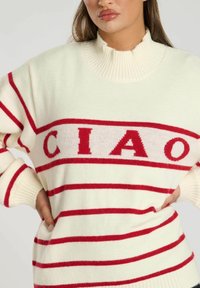 Crème trui met rode horizontale strepen en "CIAO" in vetgedrukte letters. Bevat een hoge ribgebreide halslijn en ribgebreide manchetten. Zachte textuur.