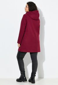 Cappotto cappuccio bordeaux con taglio dritto, texture morbida e maniche lunghe, abbinato a jeans skinny grigio scuro e stivaletti neri con lacci.
