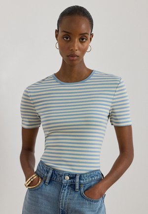 Femme à la peau foncée et aux cheveux tirés en arrière portant des boucles d'oreilles créoles en or, un t-shirt à manches courtes rayé bleu et blanc, un jean bleu et des bracelets.