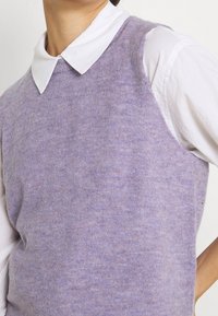 Fialový pletený svetr bez rukávů s jemnou texturou a kulatým výstřihem, nošený přes bílou košili s límečkem.