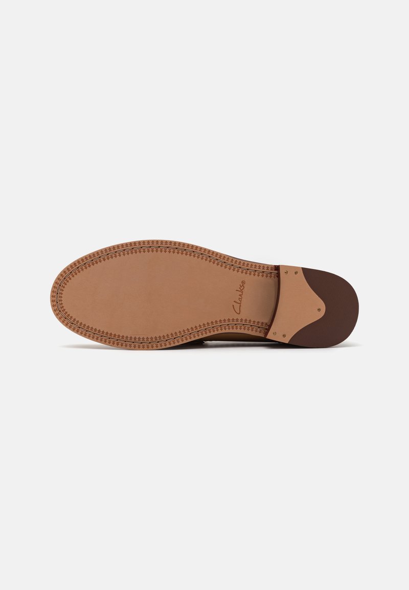 Suela de zapato de cuero color marrón claro con un borde cosido, diseño plano, textura suave y un acento de tacón de goma oscura. Marca "Clarks" visible.