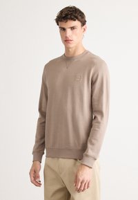 Un sweat-shirt marron à manches longues, col rond, avec un petit logo sur la poitrine. Fabriqué en tissu doux avec des poignets et un ourlet côtelés.