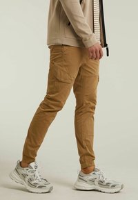 Osoba v khaki cargo kalhotách a bílých sportovních teniskách, stojící proti jednoduchému béžovému pozadí.