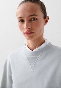 Lichtgrijze sweatshirt met een geribbelde kraag en een getextureerde driehoekige acccent. Zacht materiaal, casual ontwerp, minimalistisch en strakke lijnen.