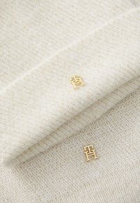 Gebreid accessoire in crème met een zachte textuur en subtiele glans. Bevat een gouden hardware logo-accent op een opgevouwen rand.