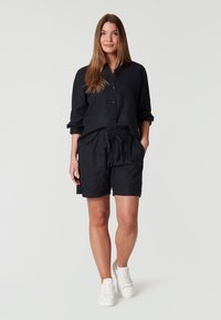 Camisa de lino negra con botones combinada con pantalones cortos negros de cordón. El modelo lleva zapatillas blancas, mostrando un estilo de atuendo casual y relajado.
