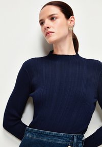 Mörkblå ribbad stickad tröja med hög krage och långa ärmar, som matchas med mörk denim, med en subtil textur och en tajt passform.