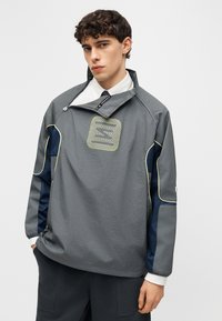 Νέος άνδρας με σγουρά μαλλιά που φοράει γκρι πουλόβερ με ναυτικό μπλε και κίτρινες λεπτομέρειες, λευκό πουκάμισο και σκούρο παντελόνι, σε απλό φόντο.