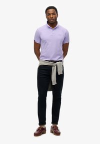 Lavendel polo shirt, medium fit, met een kraag. Grijze trui om de taille geknoopt, donkere jeans en bordeauxrode schoenen.