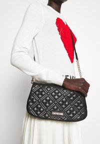 Svart kuffertväska med guldkedja och logoplack. Har en strukturerad form och en texturerad yta.