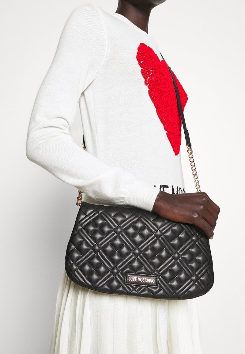 Svart kuffertväska med guldkedja och logoplack. Har en strukturerad form och en texturerad yta.