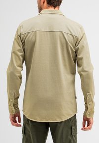 Khakifarbene Langarmbluse mit schlichter Textur, spitzem Kragen, Knopfmanschetten und abgerundetem Saum. Sichtbare Nähte und ein Etikett an der Seite.