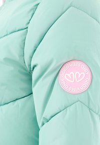 Mintgrüner, gesteppter Stoff mit einem rosa runden Patch, der zwei Herzen und den Text "NUR GUTE VIBES" am Rand zeigt.