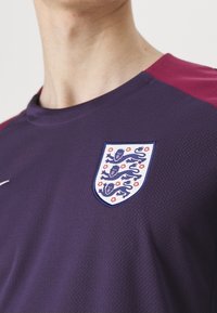 Lila sportströja i strukturerad tyg, med ett vinrött axeldesign och ett emblem av tre lejon samt röda blommor på bröstet.