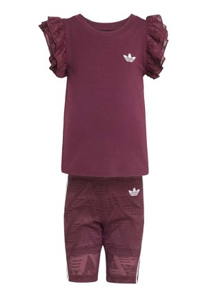 Młoda dziewczyna stoi, ubrana w bordowy top z falbanami na rękawach, dopasowane szorty, białe skarpetki w paski oraz białe tenisówki na jasnym tle.