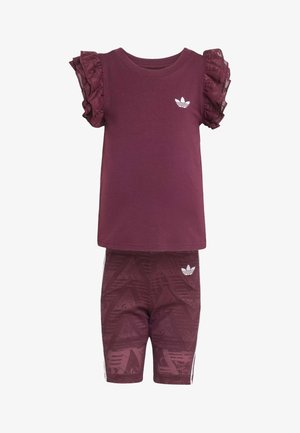 Młoda dziewczyna stoi, ubrana w bordowy top z falbanami na rękawach, dopasowane szorty, białe skarpetki w paski oraz białe tenisówki na jasnym tle.