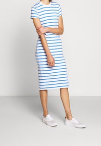 Persona che indossa un vestito midi bianco a strisce blu e sneaker bianche, in piedi davanti a uno sfondo chiaro e semplice.