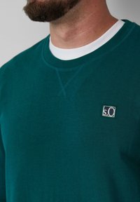 Teal sweatshirt met een ronde hals, met een witte binnenkraag. Kleine logo patch in donkerblauw en wit aan de voorkant. Zachte stofstructuur.