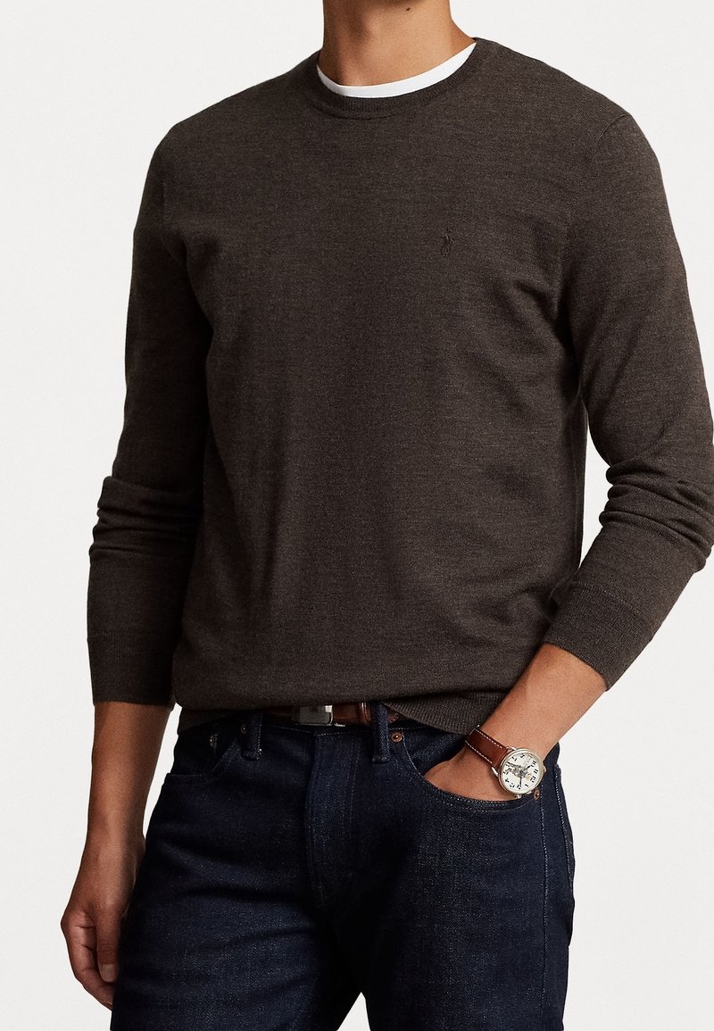 Homme portant un pull col rond à manches longues marron foncé par-dessus une chemise blanche, un jean foncé et une montre à bracelet en cuir marron avec un cadran argenté.