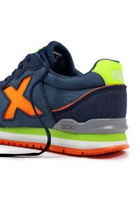 Zapatilla con parte superior azul marino, gran logotipo naranja, acentos verdes vibrantes y talón gris. Presenta un tejido texturizado y un diseño de suela en capas.