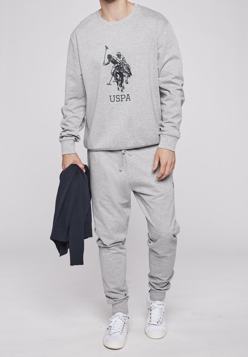 Homme en survêtement gris clair avec logo de joueur de polo et texte USPA sur le sweat-shirt, tenant un vêtement bleu marine, portant des baskets blanches.