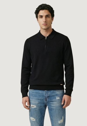 Giovane uomo con maglione nero a maniche lunghe con cerniera e colletto, e jeans chiari strappati, in piedi contro uno sfondo chiaro e uniforme.