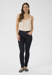 Bēšas krāsas blakus apģērba ar mežģīnēm, tumši zili skinny džinsi, melnas studētas apavi; modele stāv ar vienu roku kabatā uz vienkārša fona.