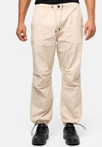 Lyse beige, elastiske joggerbukser med snoretræk og sorte hardwaredetaljer. Har sidelommer og tapered cuffs. Bæres med sorte sko.