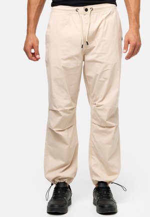 Pantalones jogger elásticos de un beige claro con cordón y detalles en negro. Presentan bolsillos laterales y puños ajustados. Se llevan con zapatos negros.