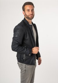 Giacca in pelle nera con zip frontale, spalle imbottite, tasche laterali e finitura testurizzata. Indossata sopra una camicia grigia chiara e jeans.