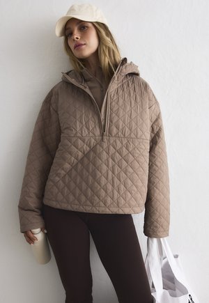 Kvinne iført beige vattert jakke og caps, holder en gjenbrukbar vannflaske og en hvit handlenettpose mot en hvit vegg.