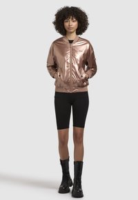 Bronzová metalická bomber bunda s žebrovanými manžetami a lemem, zapínání na zip vpředu, a dvěma bočními kapsami, nošená přes černé šortky a šněrovací boty.