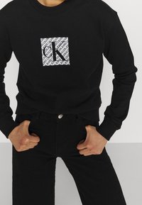 Sudadera negra con un logo cuadrado que presenta un patrón repetido y la letra 'K'. Combinada con jeans negros. Diseño simple y casual.