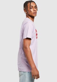Lilla T-shirt med korte ærmer, der har fed rød tekst. T-shirten har en afslappet pasform med rund hals og er lavet af blødt bomuldsmateriale.