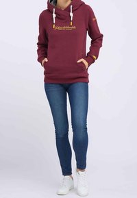 Sudadera con capucha color burdeos y cuello tipo cowl, con acentos amarillos, bolsillos frontales y texto bordado. Combinada con jeans azules y zapatillas blancas.