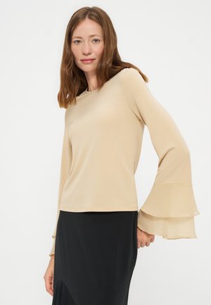 Kvinde med glat brunt hår bærer beige langærmet top med lagdelte klokkeærmer og et sort skørt mod ensfarvet baggrund.
