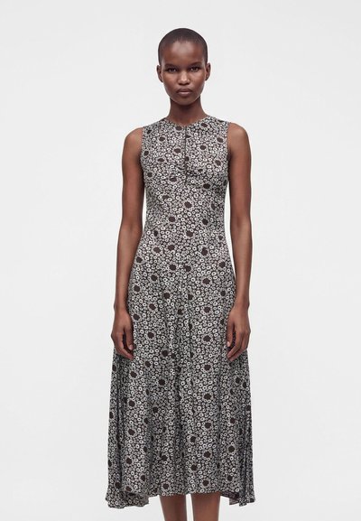 Femme portant une robe noire sans manches avec un petit imprimé floral blanc, debout droite sur un fond uni clair.
