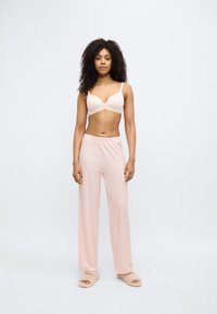 Light pink bra with soft fabric, V-neck design. Flowing light pink pajama pants with subtle pattern, paired with beige fuzzy slippers.