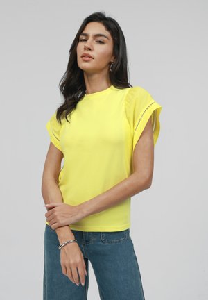 Chemise jaune à manches courtes avec un col rond et des manches froncées. Le tissu semble lisse avec des découpes latérales subtiles. Se porte avec un jean bleu.