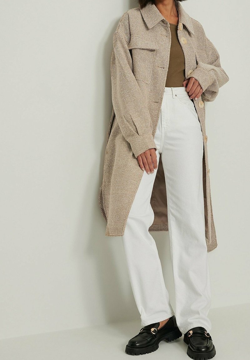 Beige, getextureerde lange jas met twee voorzakken, gecombineerd met witte jeans en zwarte loafers met een gouden accent.