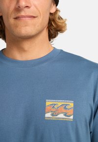 Blå bomull T-skjorte med en fargerik bølgedesign i oransje og gult innenfor en rektangulær kontur, med ribbestrikket rund hals.