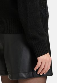 Zwarte gebreide trui met geribbelde manchetten, gecombineerd met glanzende zwarte leren shorts. De texturen contrasteren tussen zachte wol en slank leer.