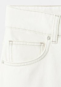 Détail d'un jean en denim blanc montrant la poche avant, la bride de ceinture et les rivets métalliques avec une couture marron clair.