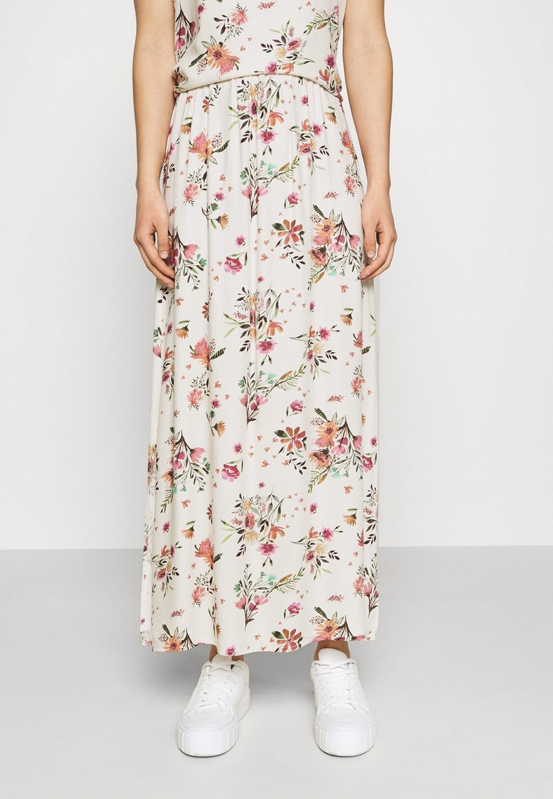 Femme portant une jupe longue blanche à fleurs roses et vertes, associée à des baskets blanches à plateforme, debout devant un fond uni.